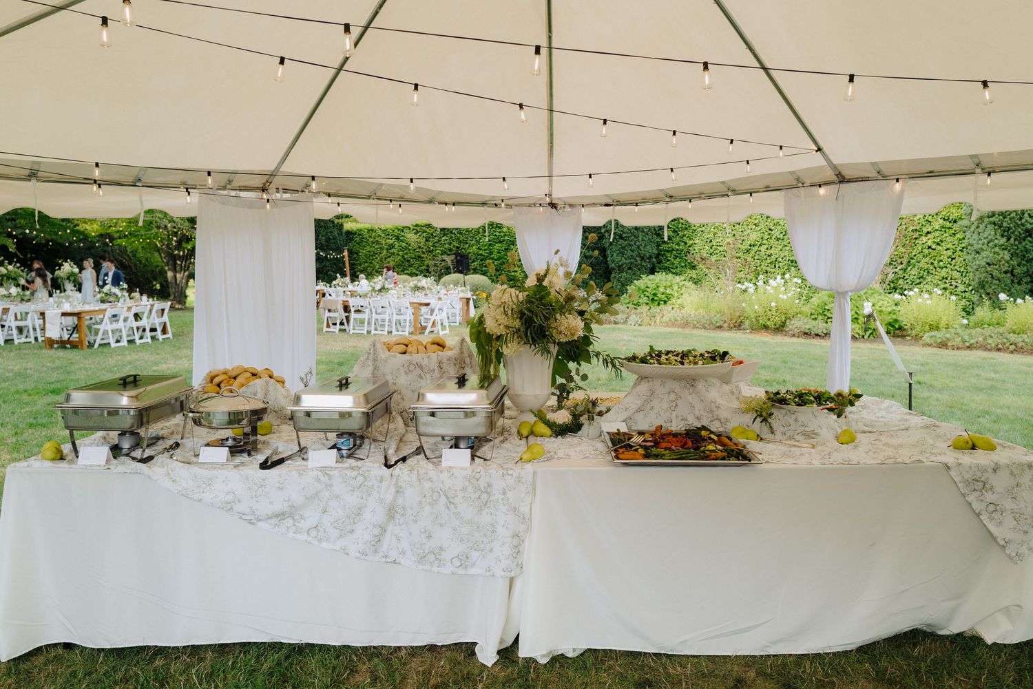 A white tent with buffet tables and chafing dishes set up for an outdoor wedding reception.