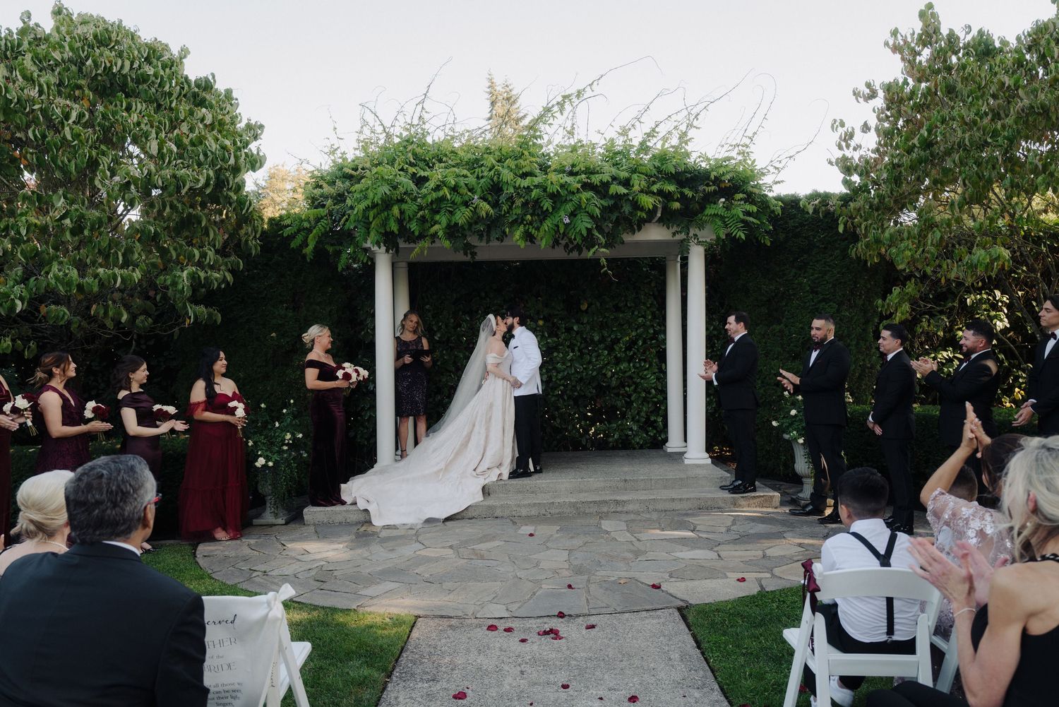 Outdoor wedding ceremony setup with white chairs and floral arrangements in a garden setting.
