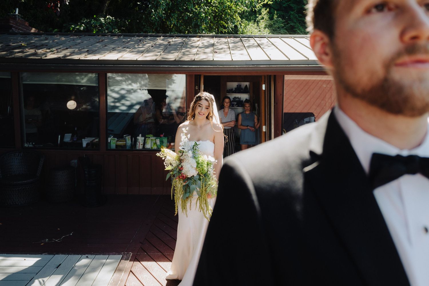 A wedding couple walks together in formal attire through a sunny outdoor venue with decorative lighting.