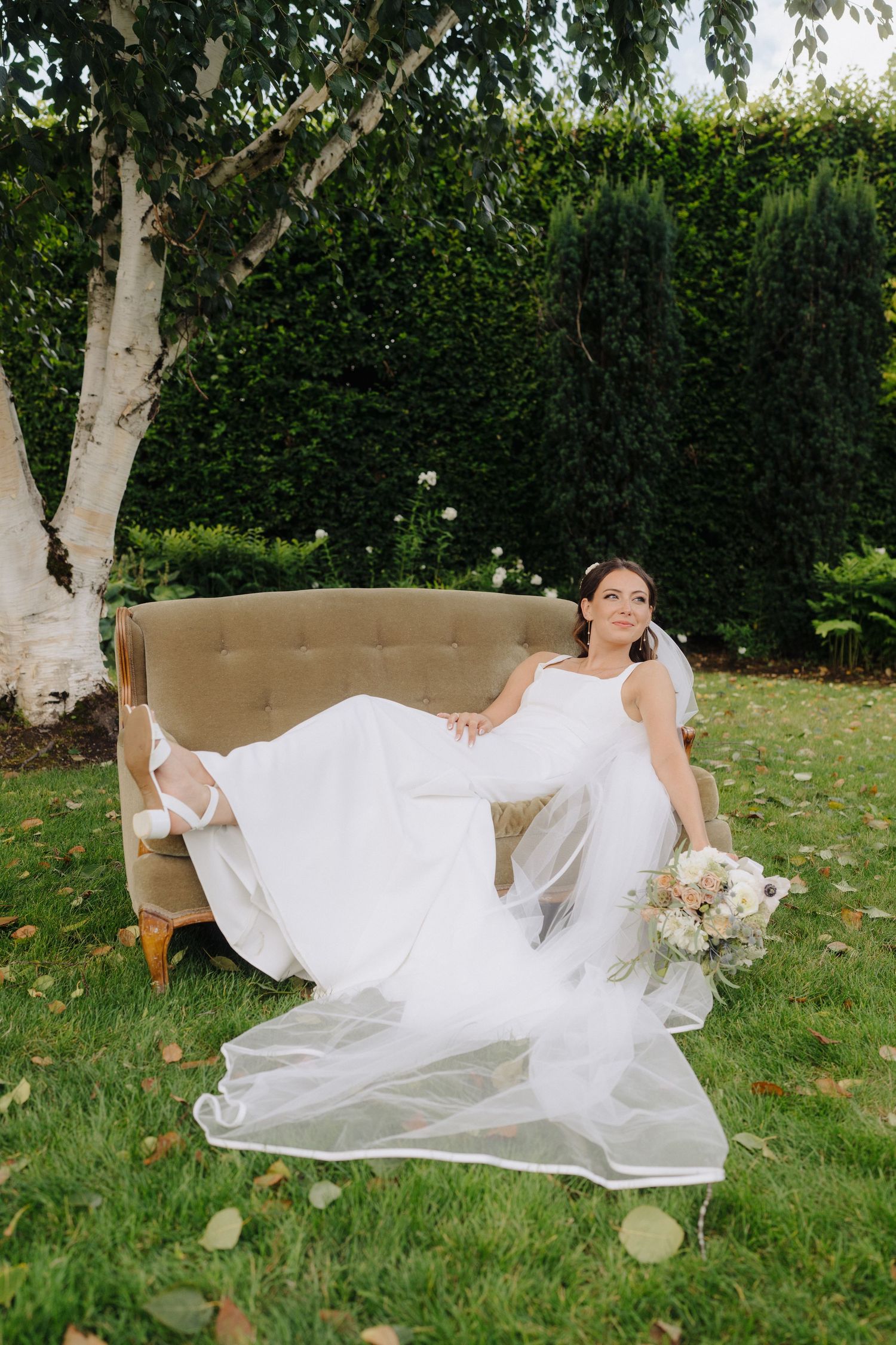 A white wedding dress flows elegantly across a grassy lawn with lush green hedges in the background.