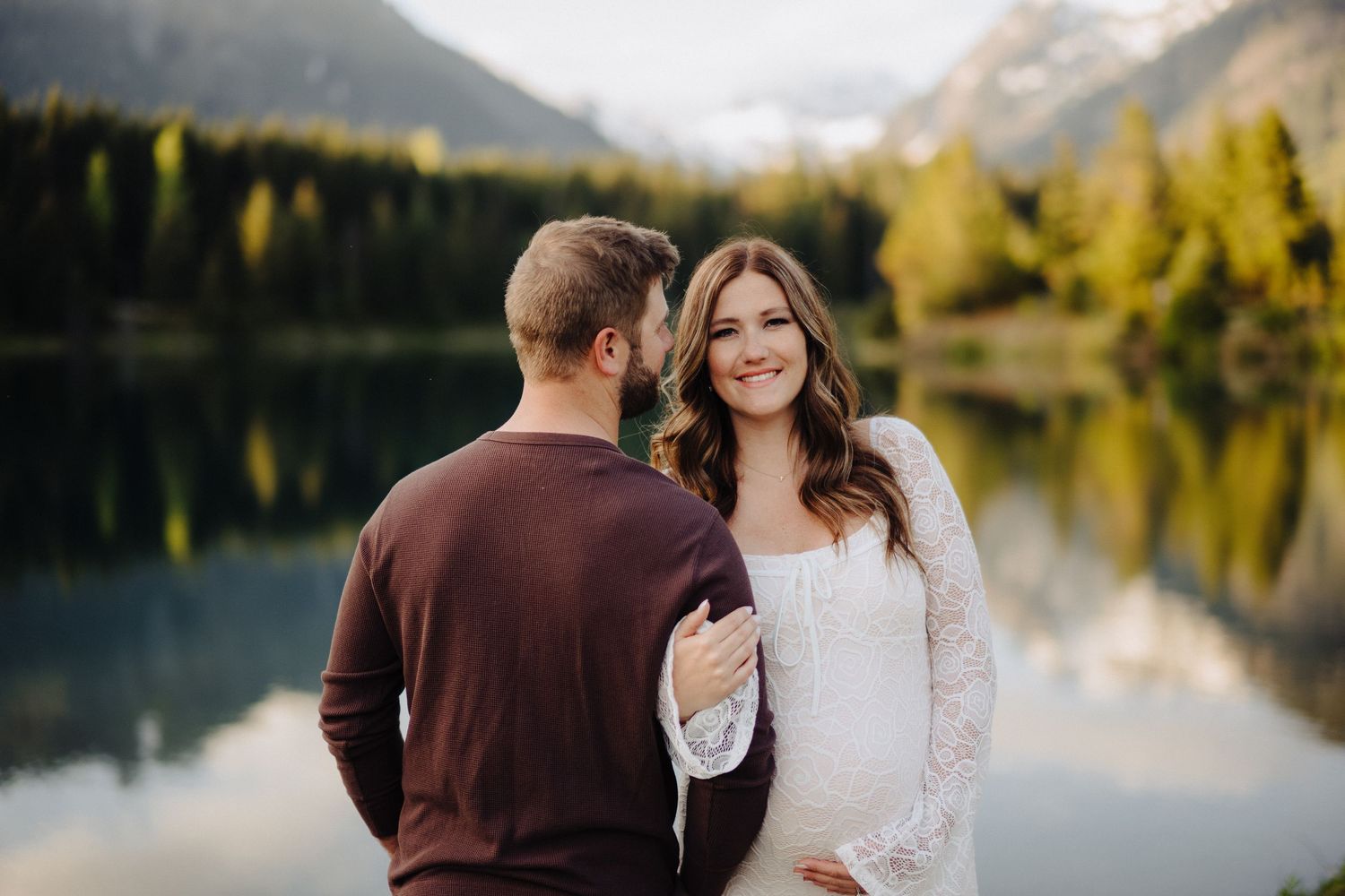 Romantic lakeside engagement photos with mountain backdrop and golden autumn trees reflecting in calm waters.