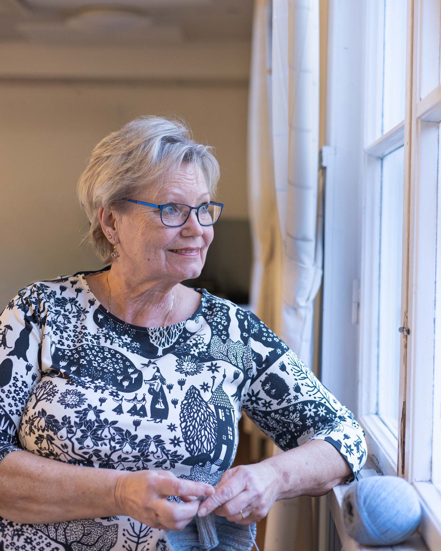 Värikuva ikkunan äärellä istuvasta ja neulovasta iäkkäämmästä naisesta. Potrettikuvaus Tuusulassa.