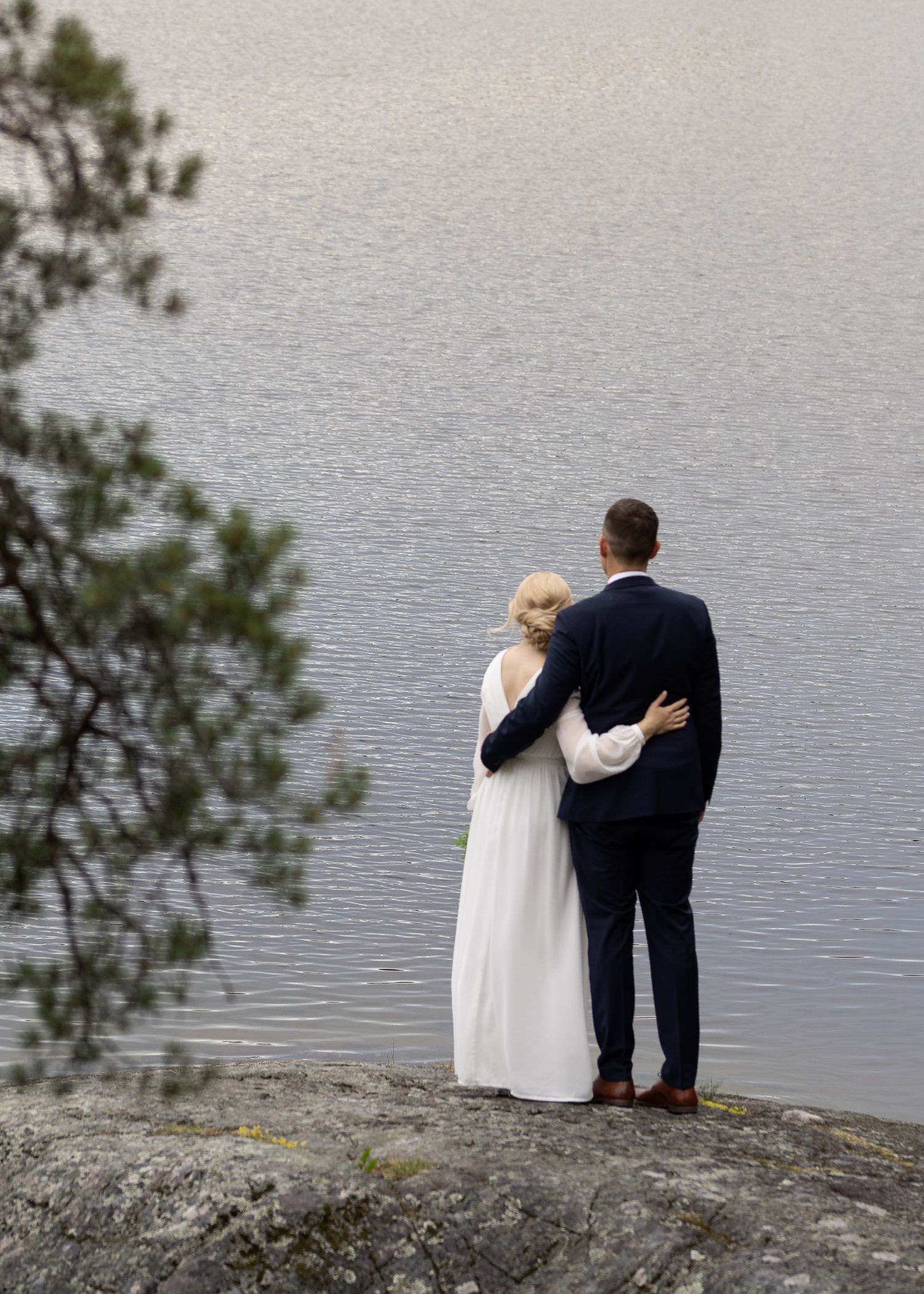 Hääpari seisoo lähekkäin korkealla kalliolla suuren järvimaiseman äärellä. Juhlava hääkuvaus ja hääpotretit miljöössä.
