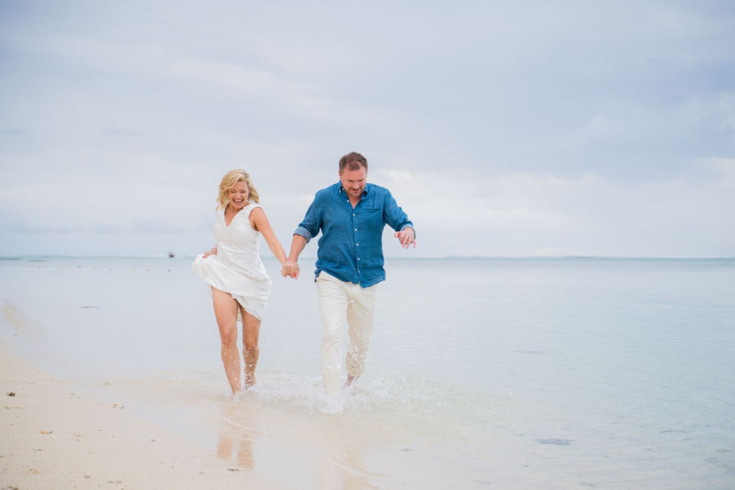 Couple holding hands running along a tropical white sand beach with calm turquoise water in the background.