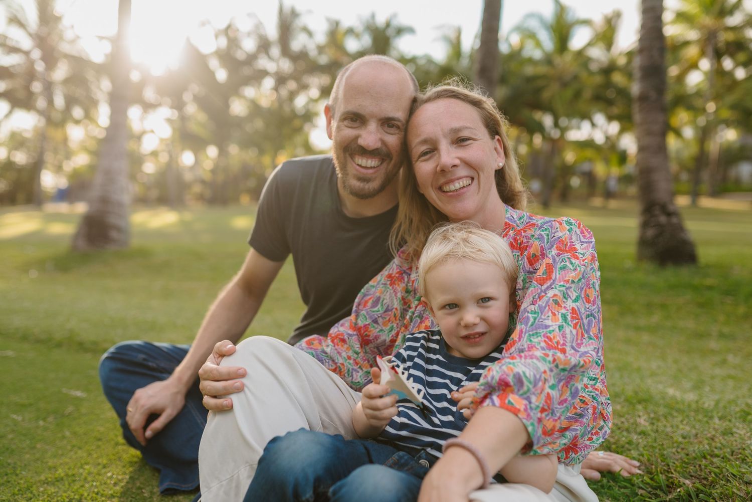 Happy family of three sitting on green grass with palm trees, parents smiling with toddler son in sunny tropical park.