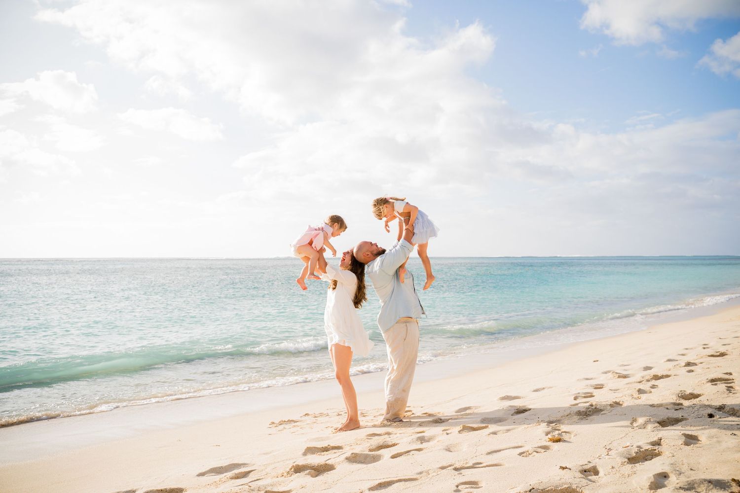 Family walking on a sunny beach, parents lifting toddlers on their shoulders, white sand and turquoise ocean backdrop.