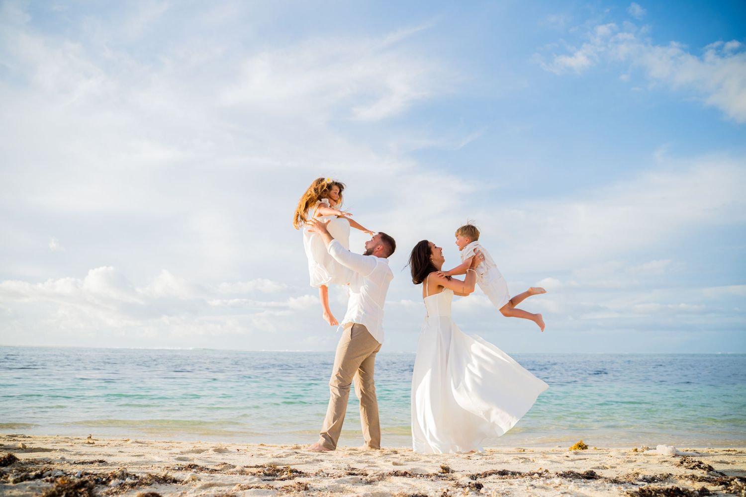 Happy family in white outfits playing on a tropical beach, parents lifting children against a blue sky.