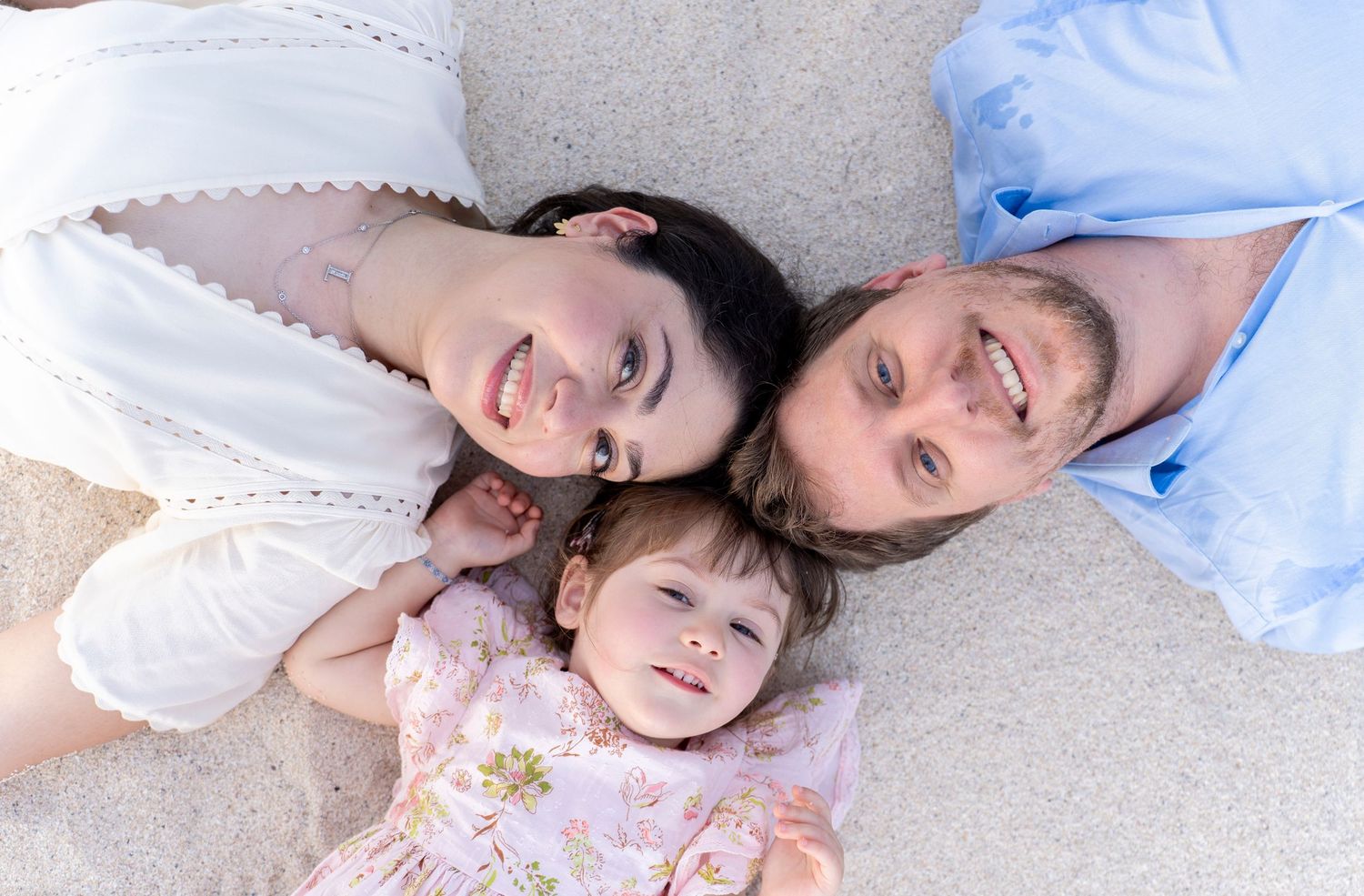 A happy family of three lying on sand, parents smiling with baby girl between them, aerial view.