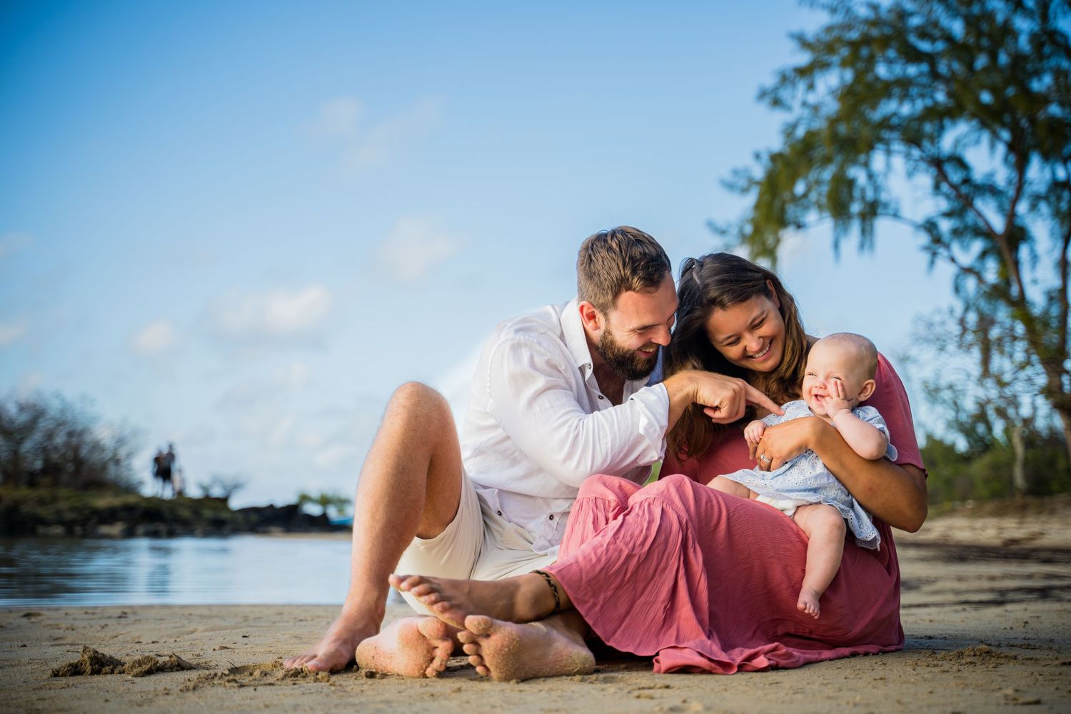 Family with baby sitting on sandy beach, parents smiling at newborn, tropical trees and blue sky background.