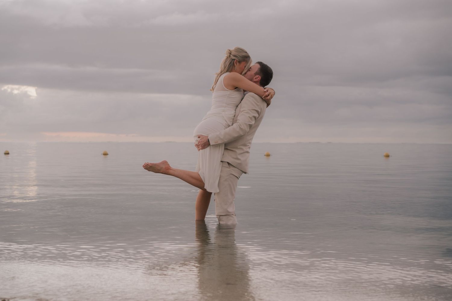 Couple embracing and kissing on a calm beach shore at sunset with cloudy skies in the background.