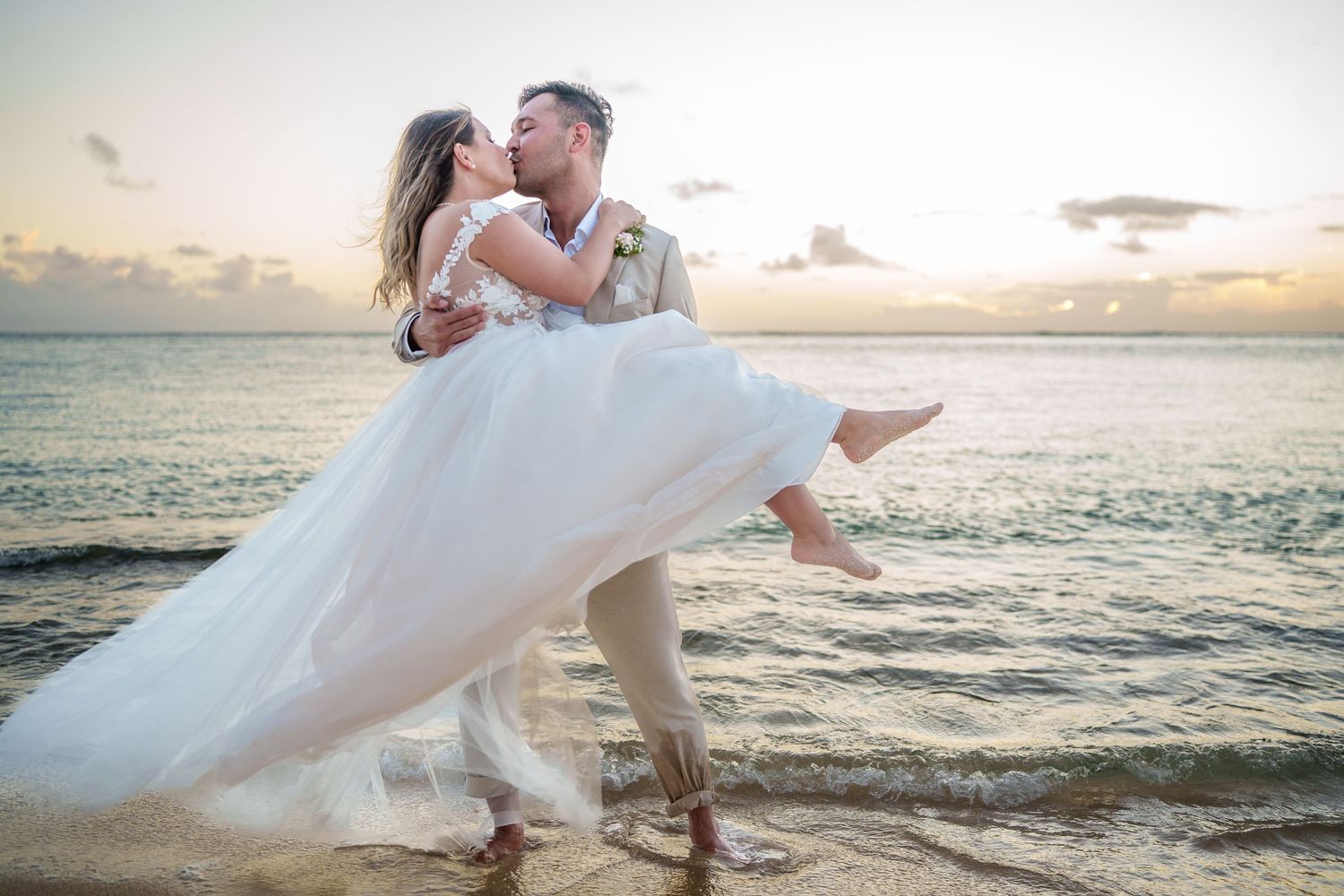 Groom lifting bride in white gown at beach sunset, sharing romantic kiss by ocean waves.