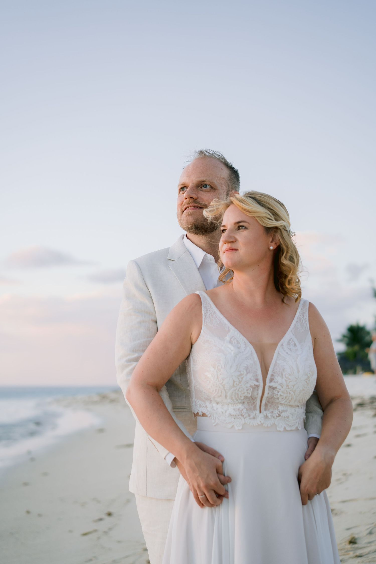 Bride and groom in white attire embrace on a sandy beach at sunset, gazing into the distance.