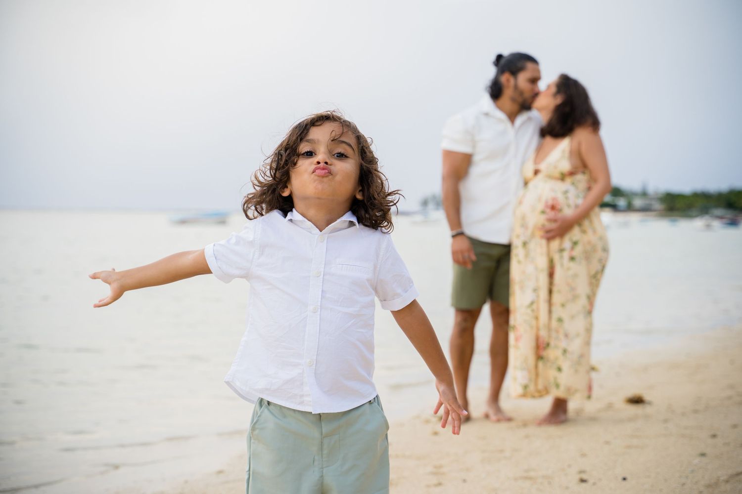 A young boy poses on a beach with arms outstretched while his pregnant mother and father kiss behind him.