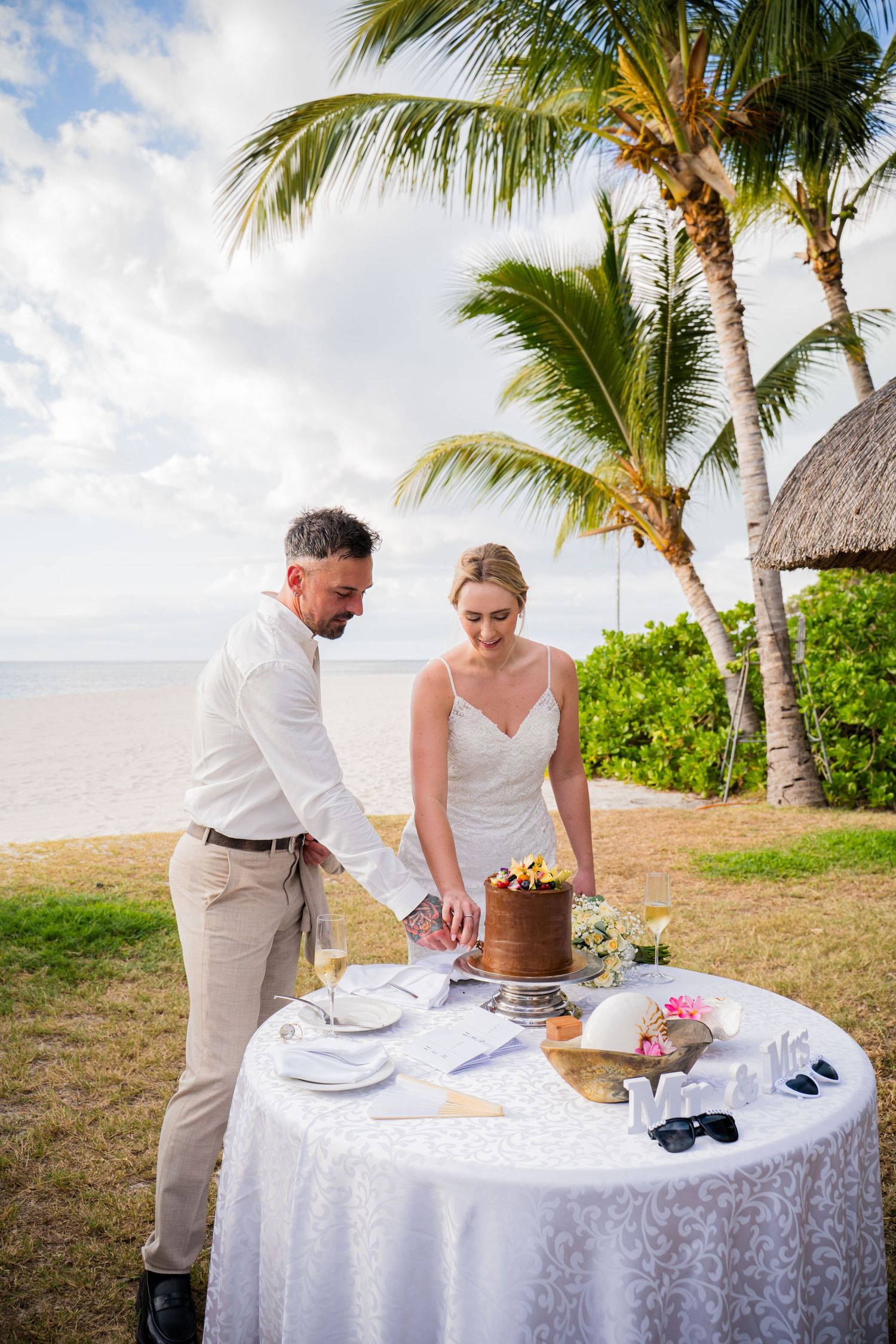 Bride and groom cutting wedding cake on tropical beach with palm trees and ocean backdrop.