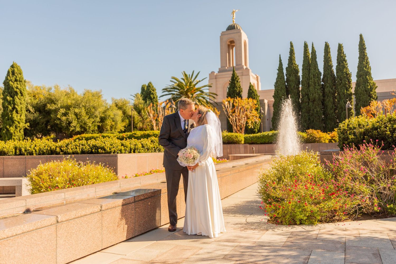 Wedding couple poses by fountain with Mediterranean church architecture in background.