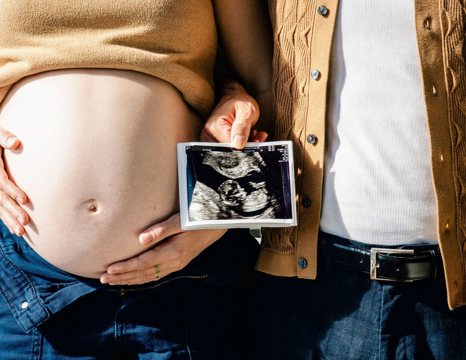 Hands hold an ultrasound photo against a pregnant belly wearing a tan sweater.