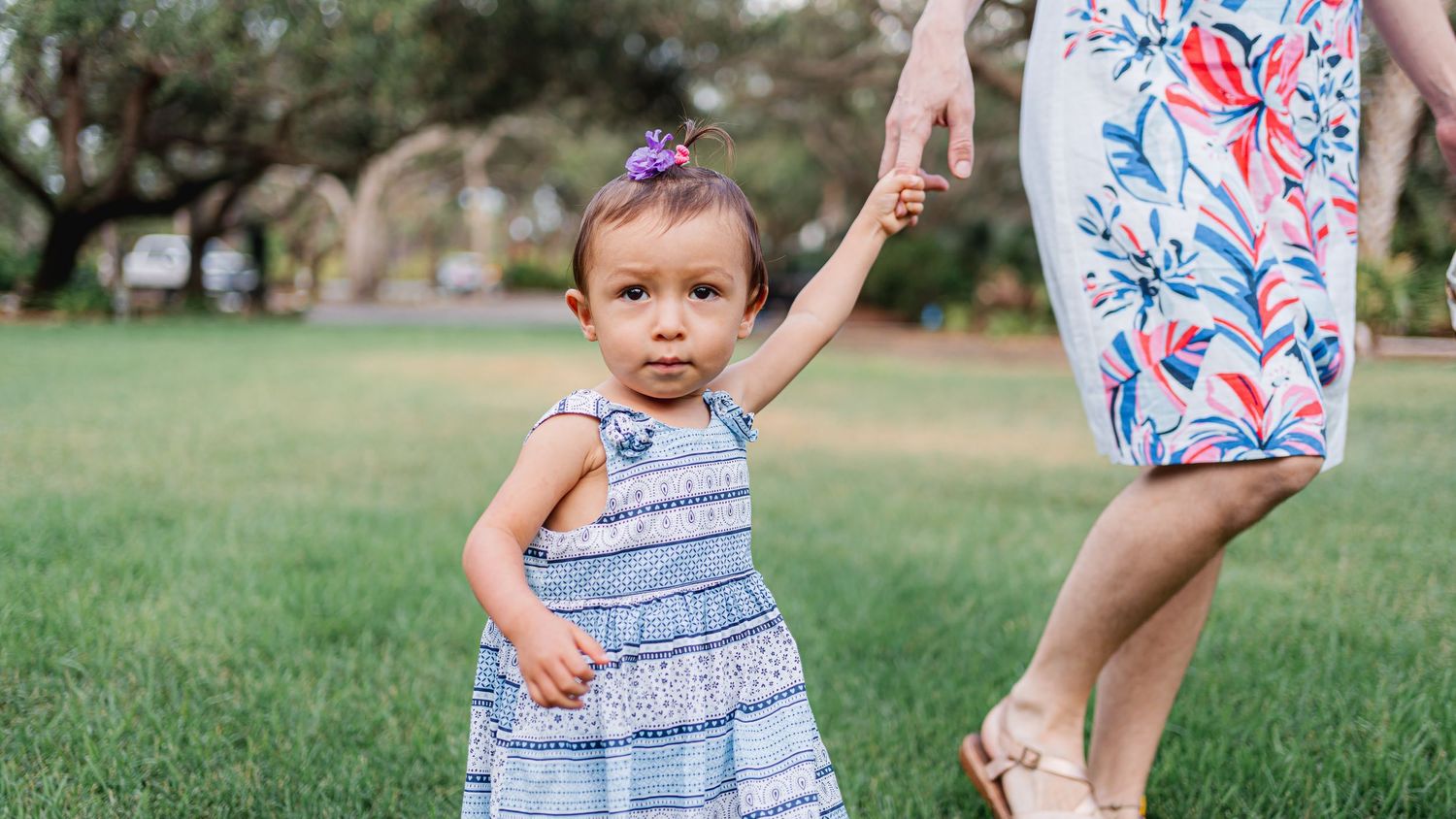 Young child in striped dress walks on grass while holding someone's hand.