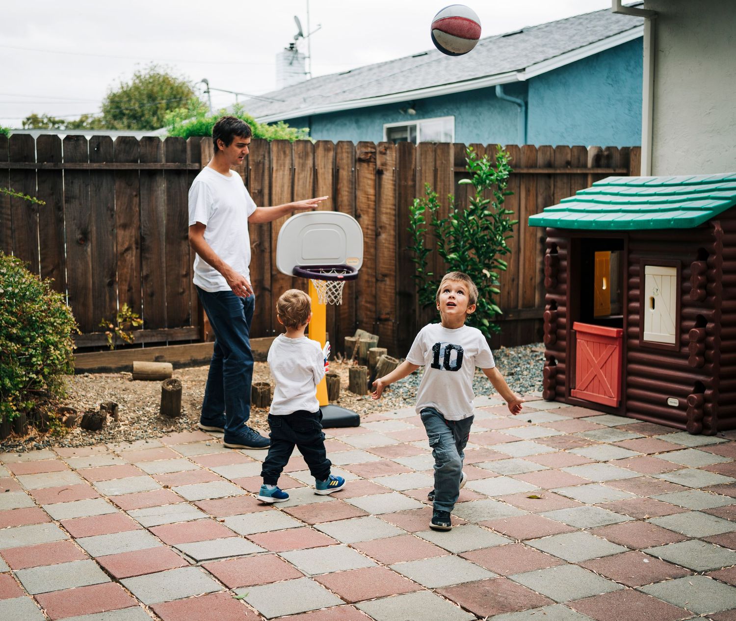 Children playing basketball and running around on a brick patio in a backyard setting.