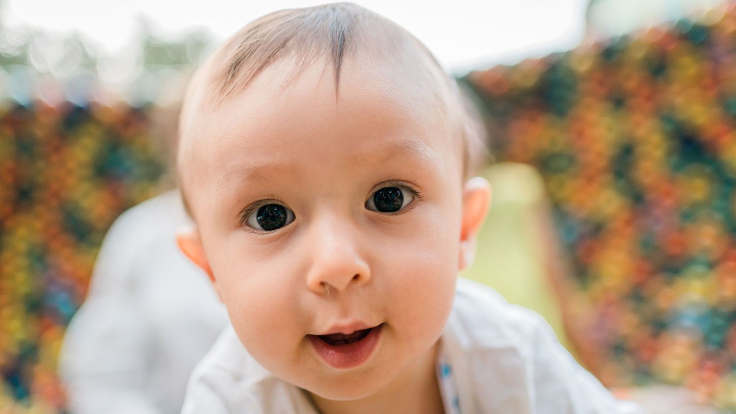 Baby with bright wide eyes and surprised expression looks at camera against colorful background.
