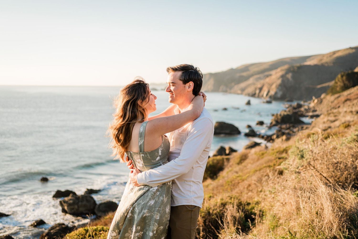 A couple embraces on a scenic cliff overlooking the Pacific Coast during golden hour at sunset.