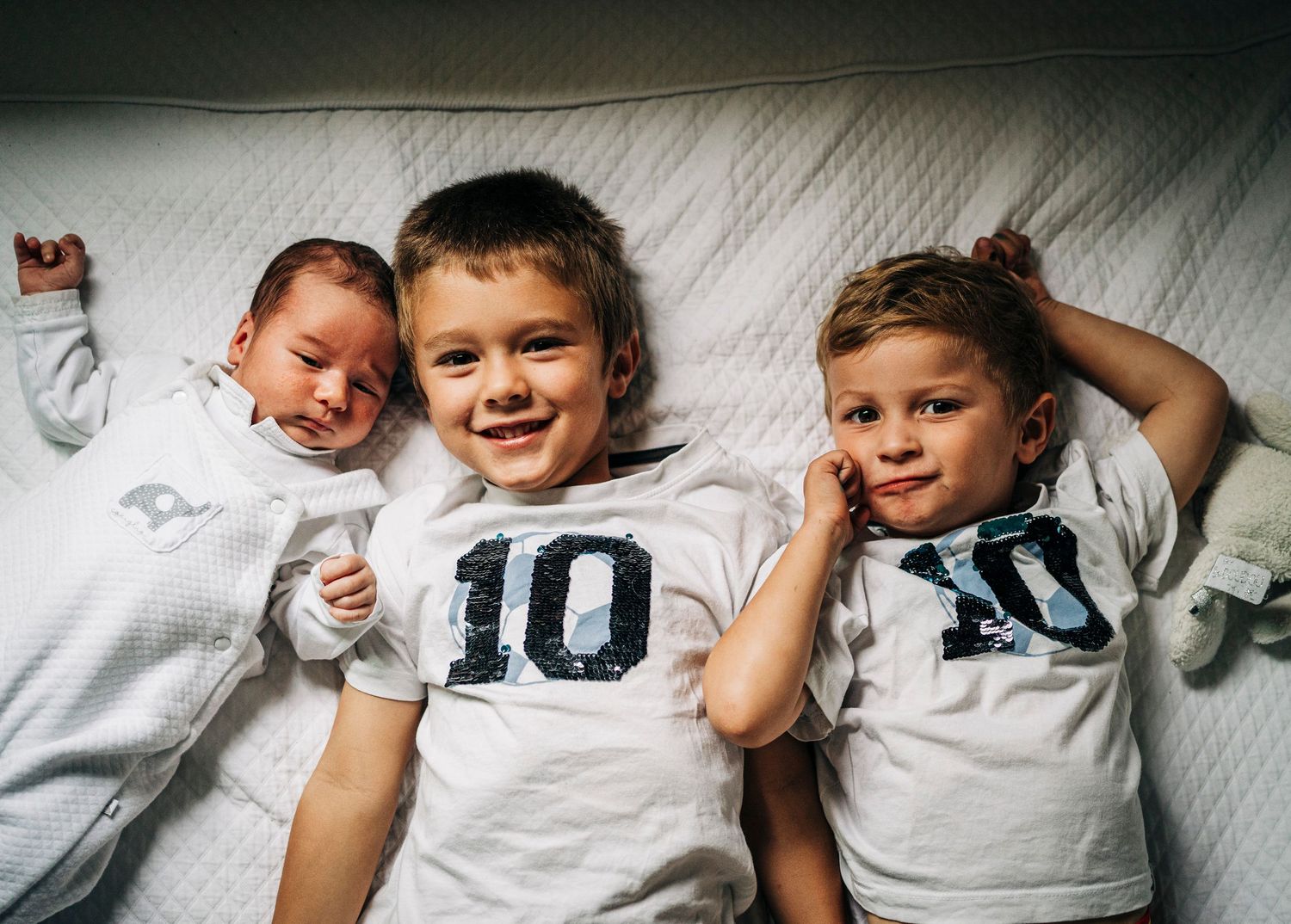 Three children in white shirts lie together on a light colored surface, playfully posing for the camera.
