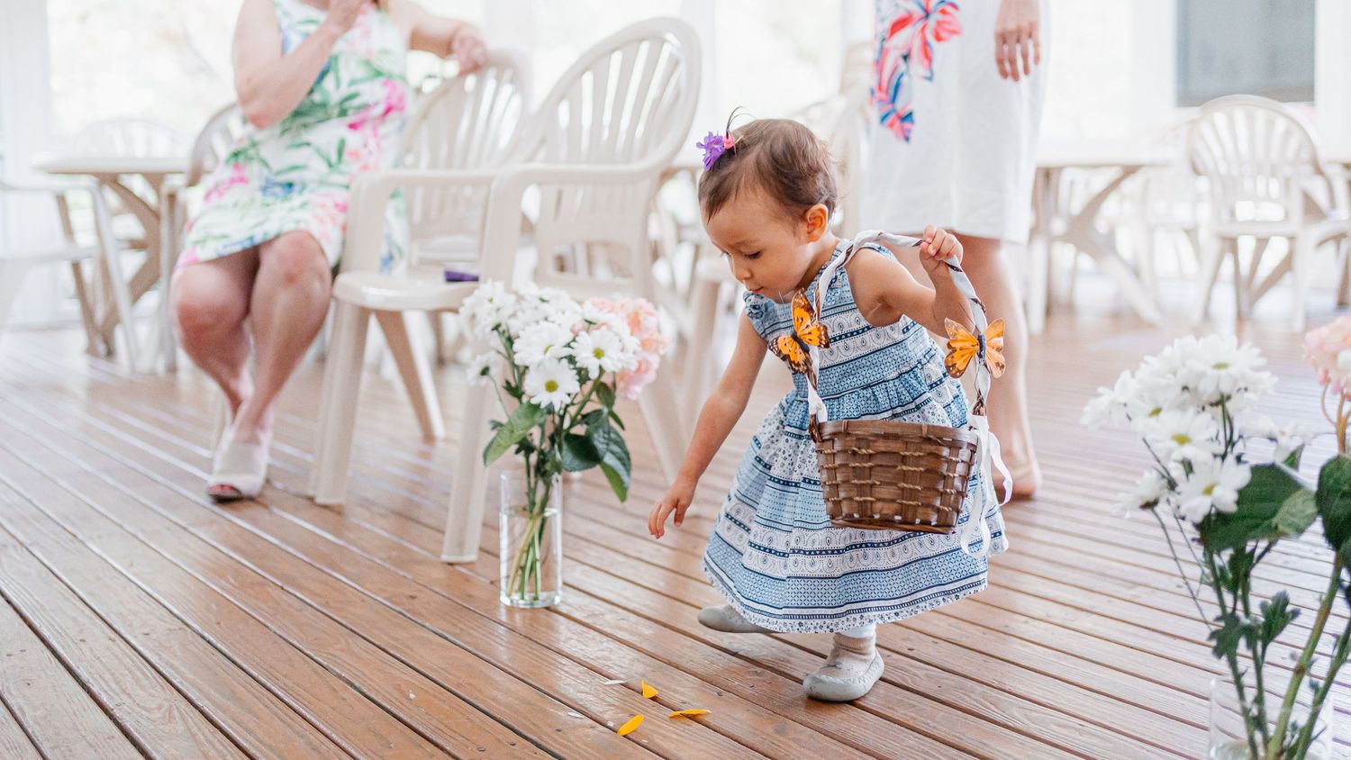 Small child in a blue dress plays on wooden deck decorated with white flowers in vases.