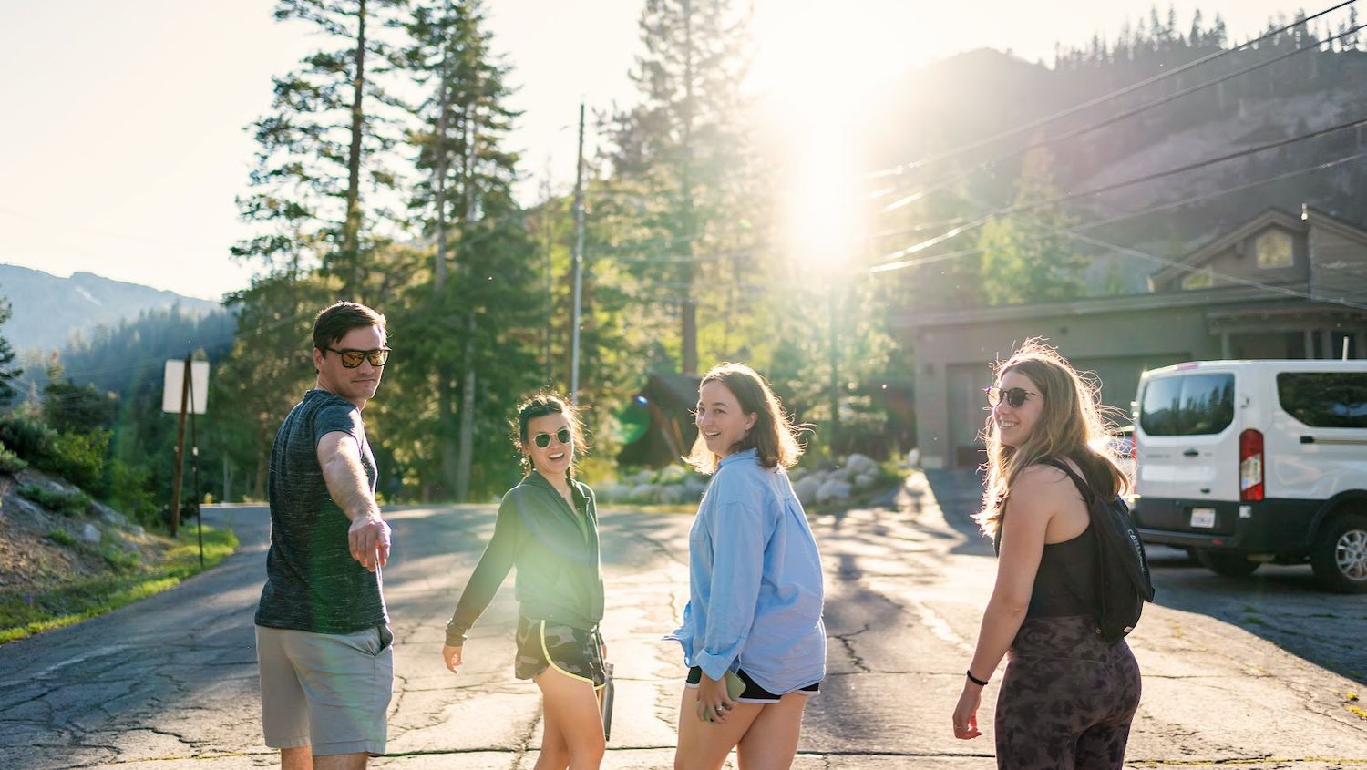 People walking along a mountain path at golden hour with sun flare through trees.
