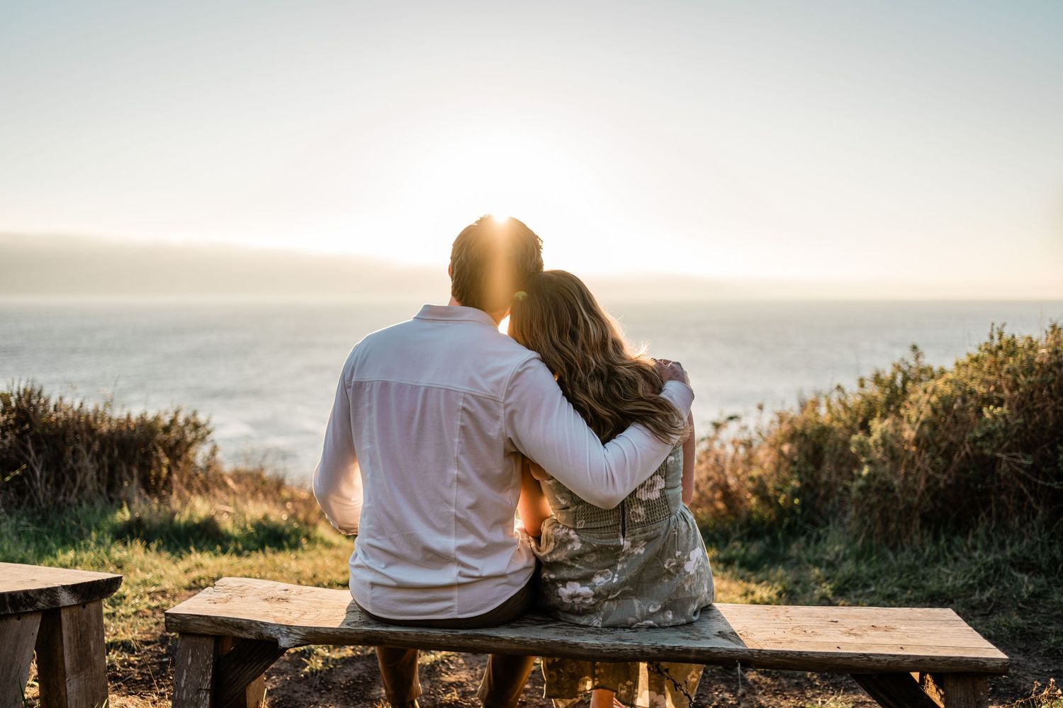 Couple sitting on a bench embracing while watching a golden sunset over the ocean from a coastal clifftop.