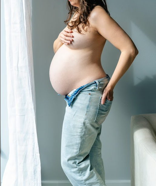 A pregnant woman in light jeans and beige top stands in profile against a gray wall by a white curtain.