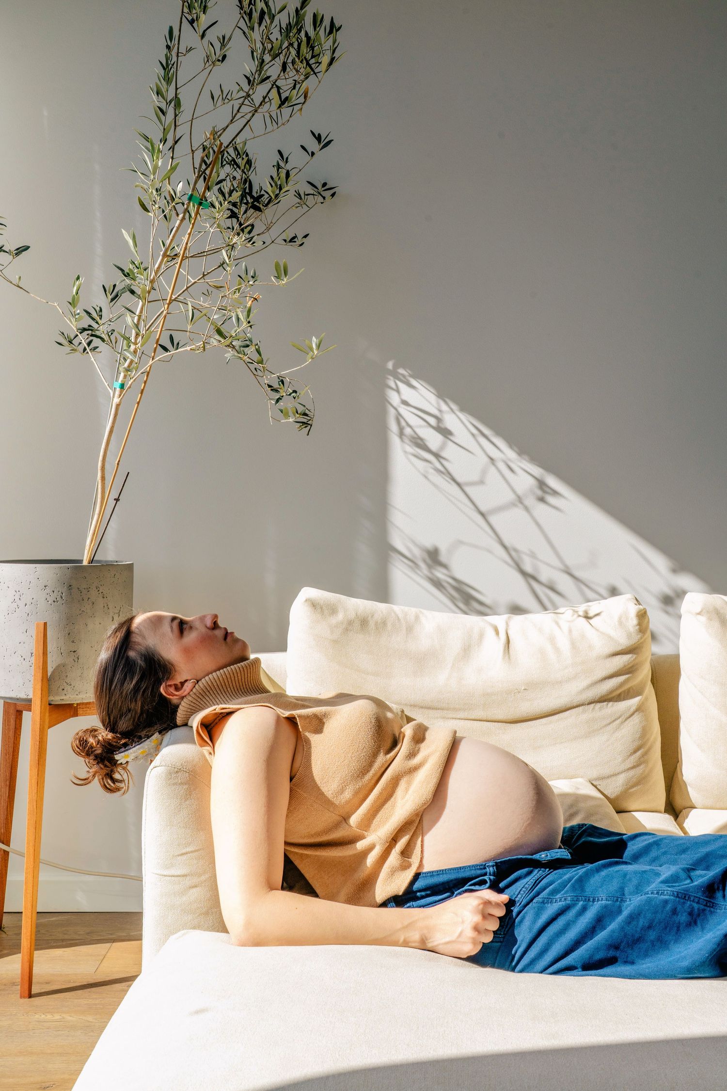 Side view of pregnant person reclining on couch near potted plant in sunlit room.
