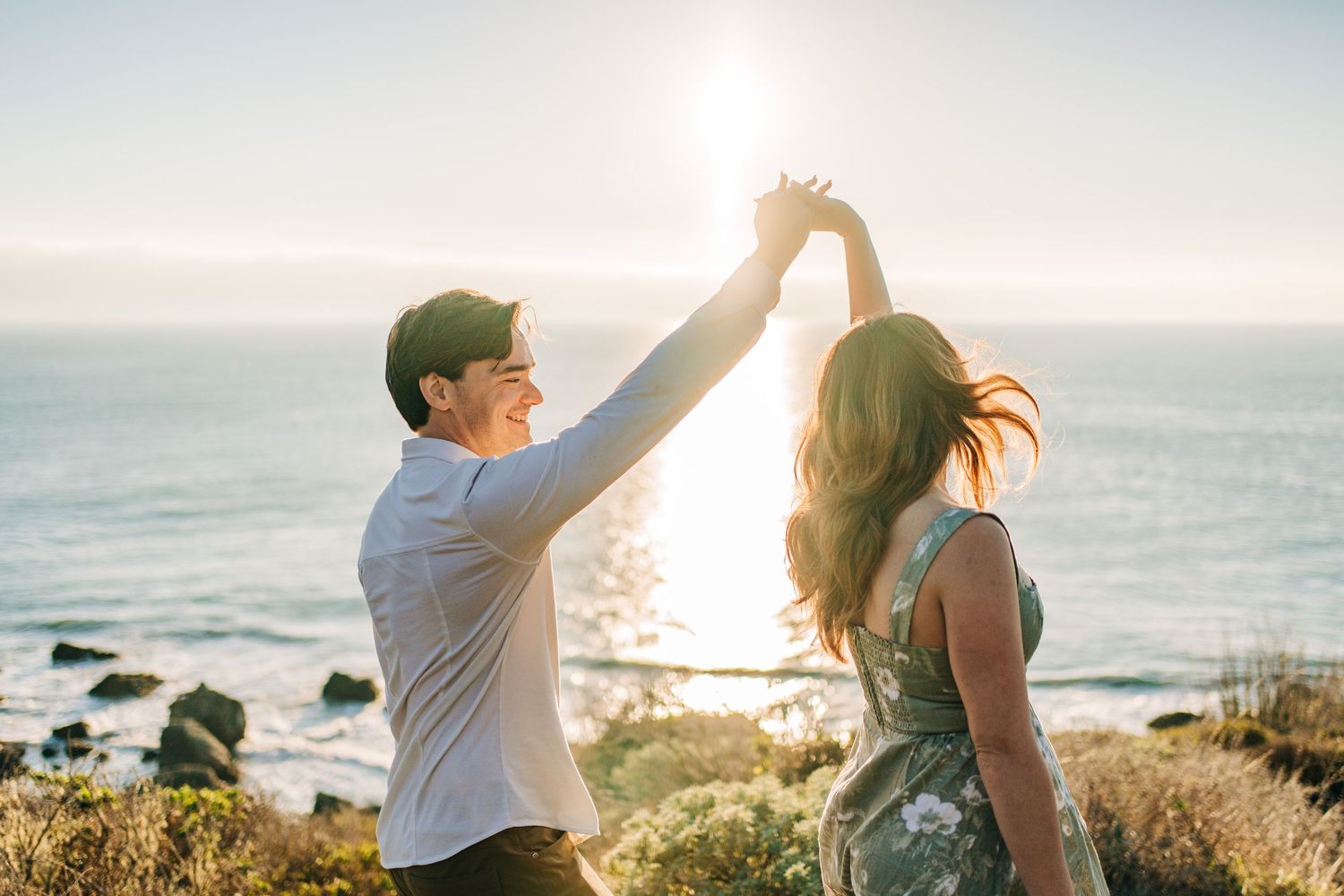 A couple dances together on a coastal cliff during golden hour with the ocean and sunlight in the background.