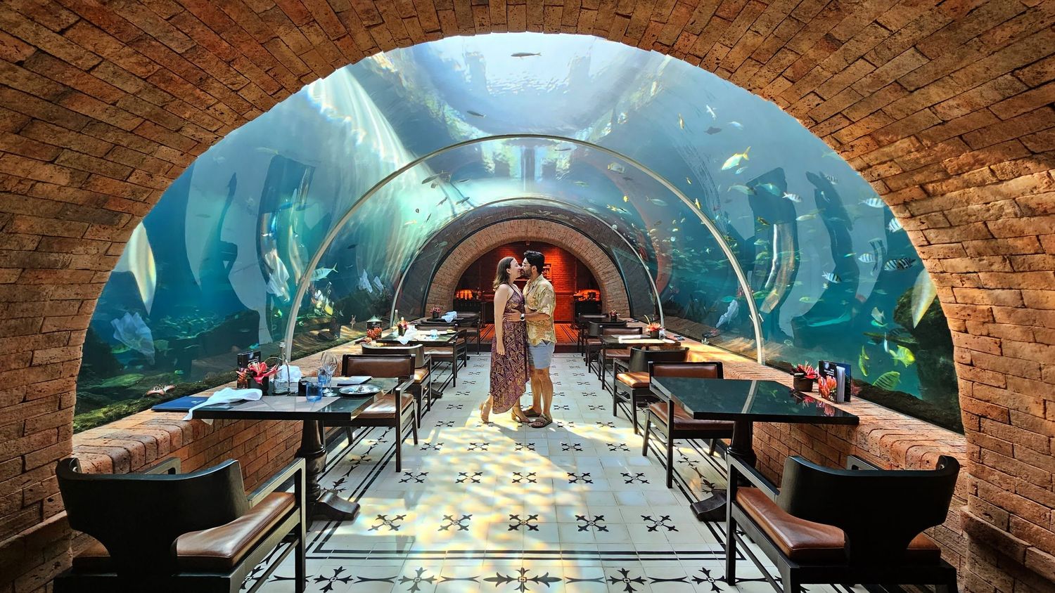 Panoramic view of curved brick tunnel with aquarium above and lighting and reflective floor.