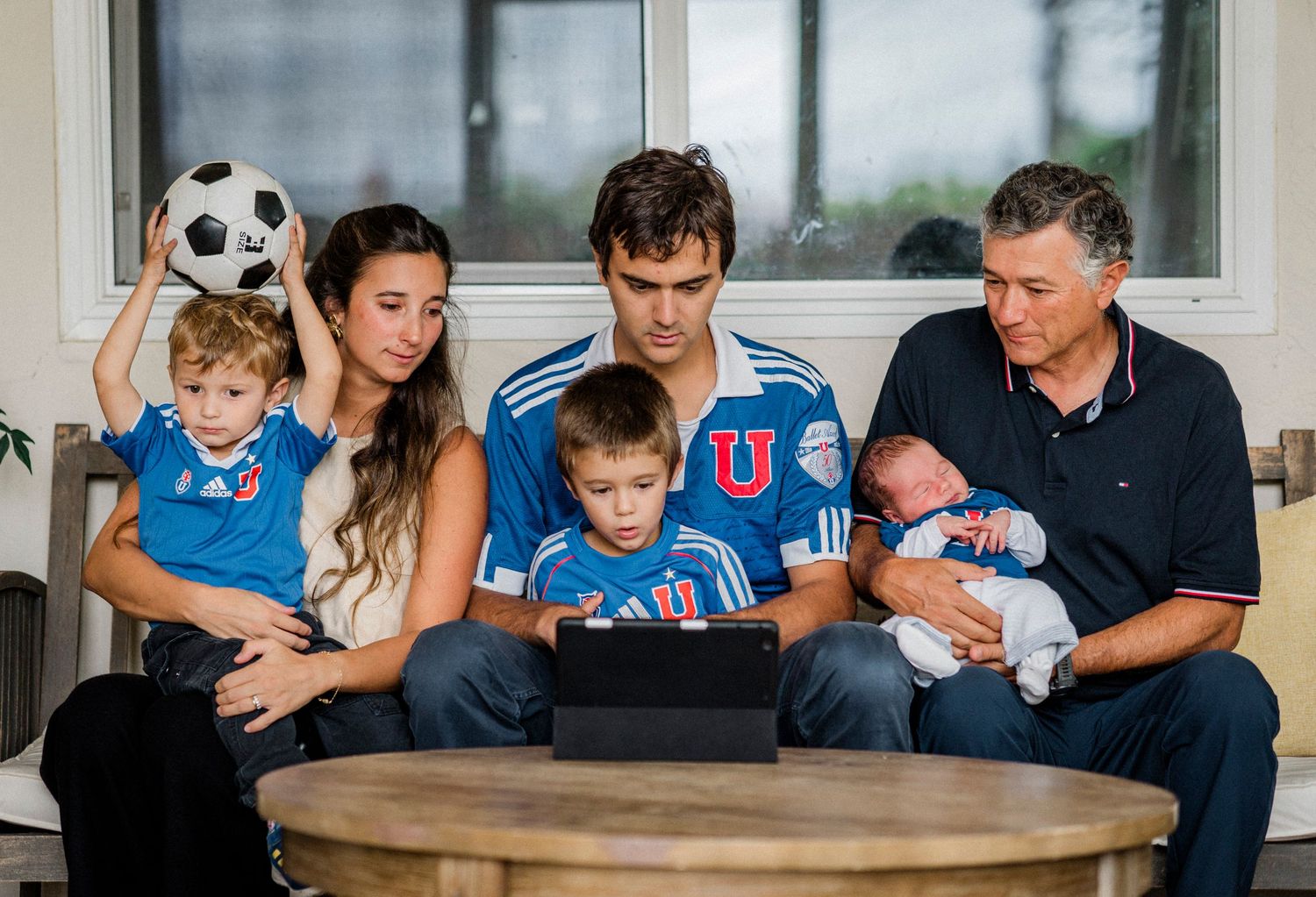 Family sitting on couch watching soccer match and playing with soccer balls in living room.