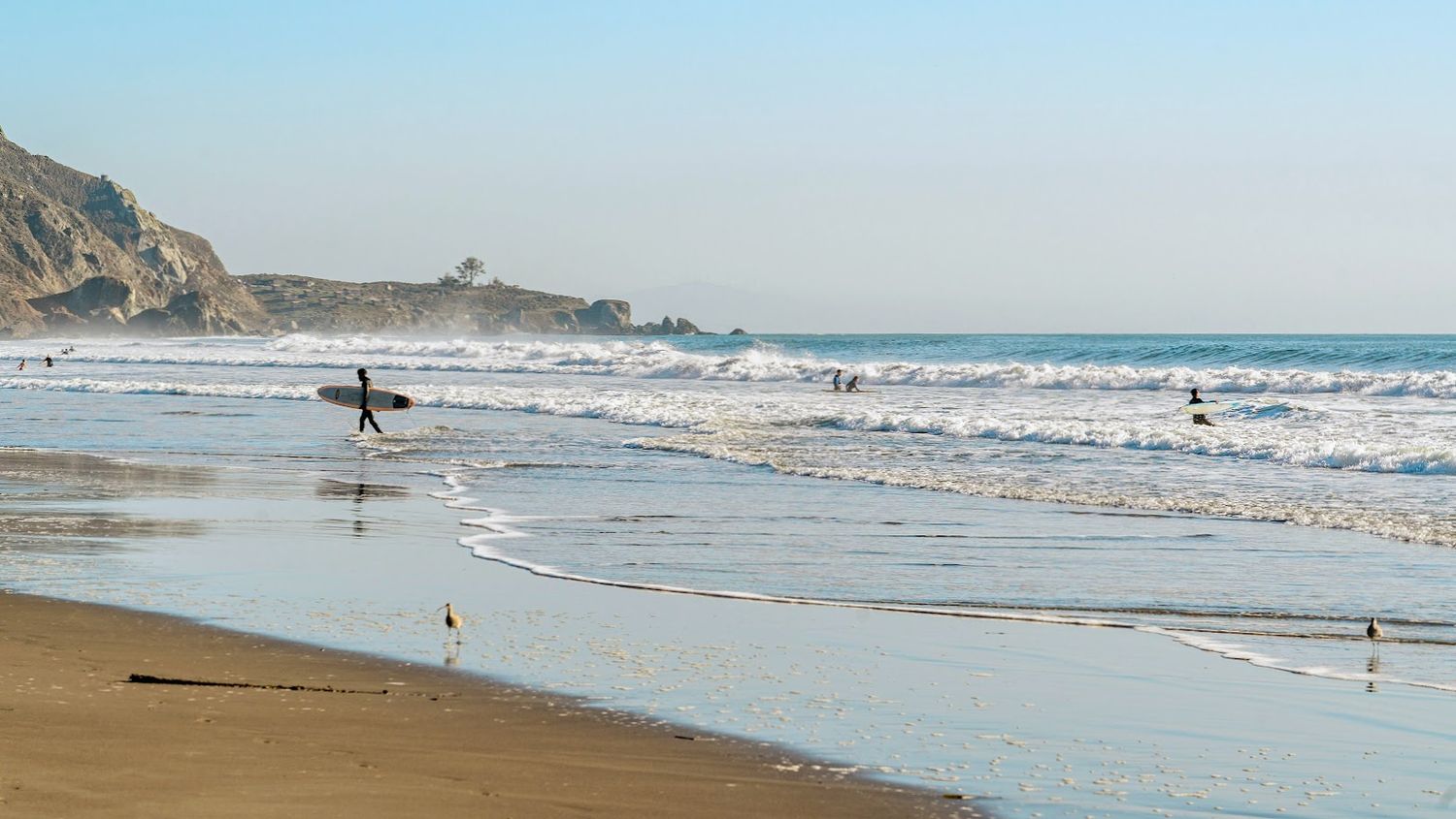 Wide sandy beach with waves rolling onto shore and coastal cliffs in the distance.