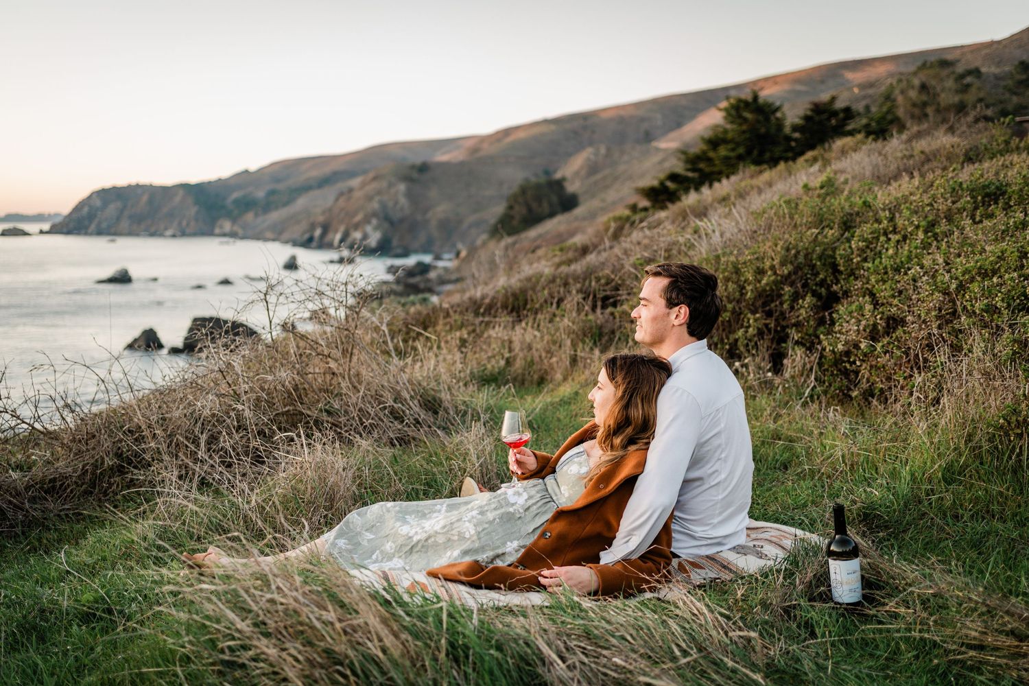 Couple enjoying wine and picnic on coastal cliffside overlooking dramatic Pacific Ocean landscape at sunset.
