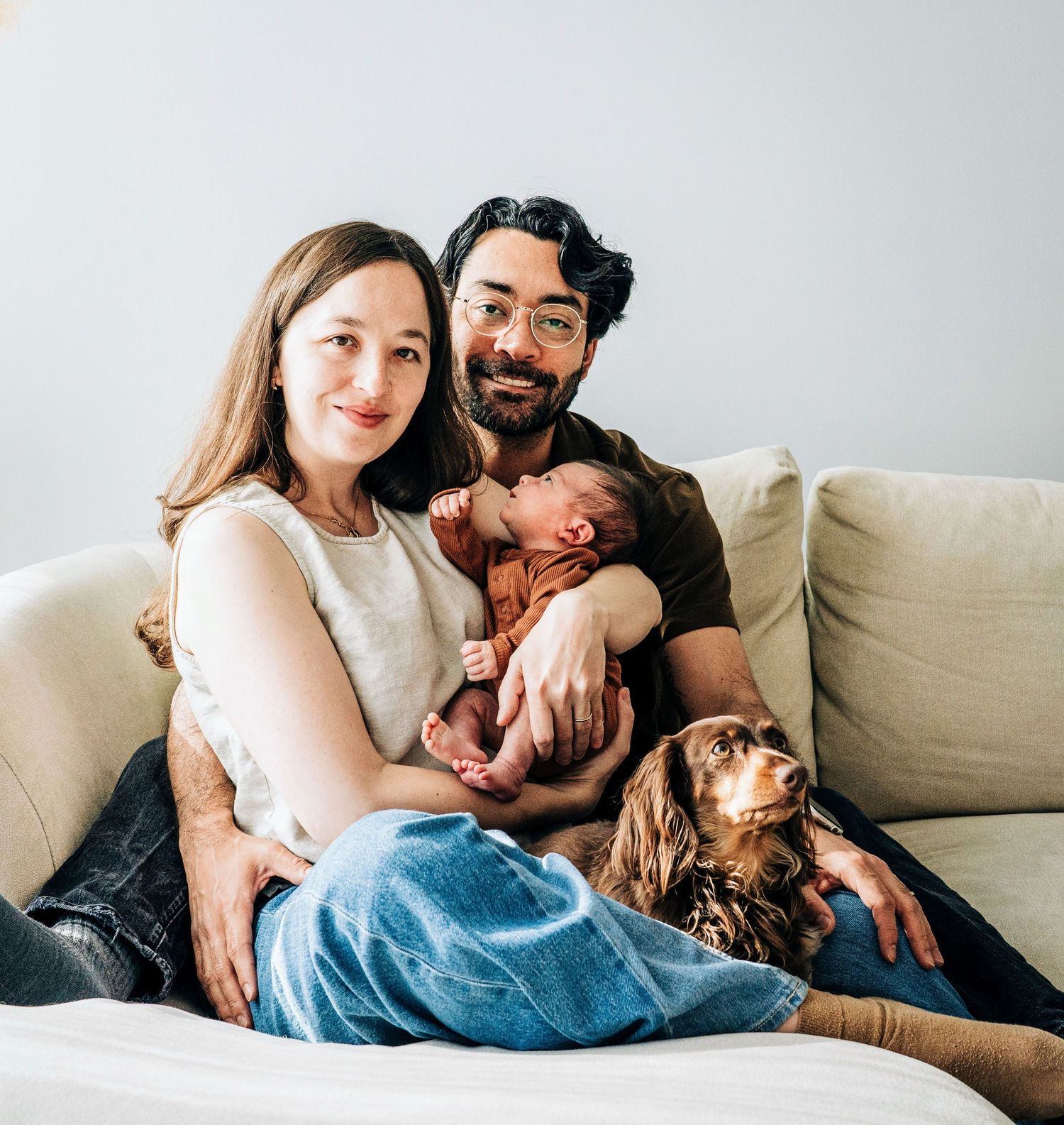 A family cuddles together on their couch with their small baby and dog.