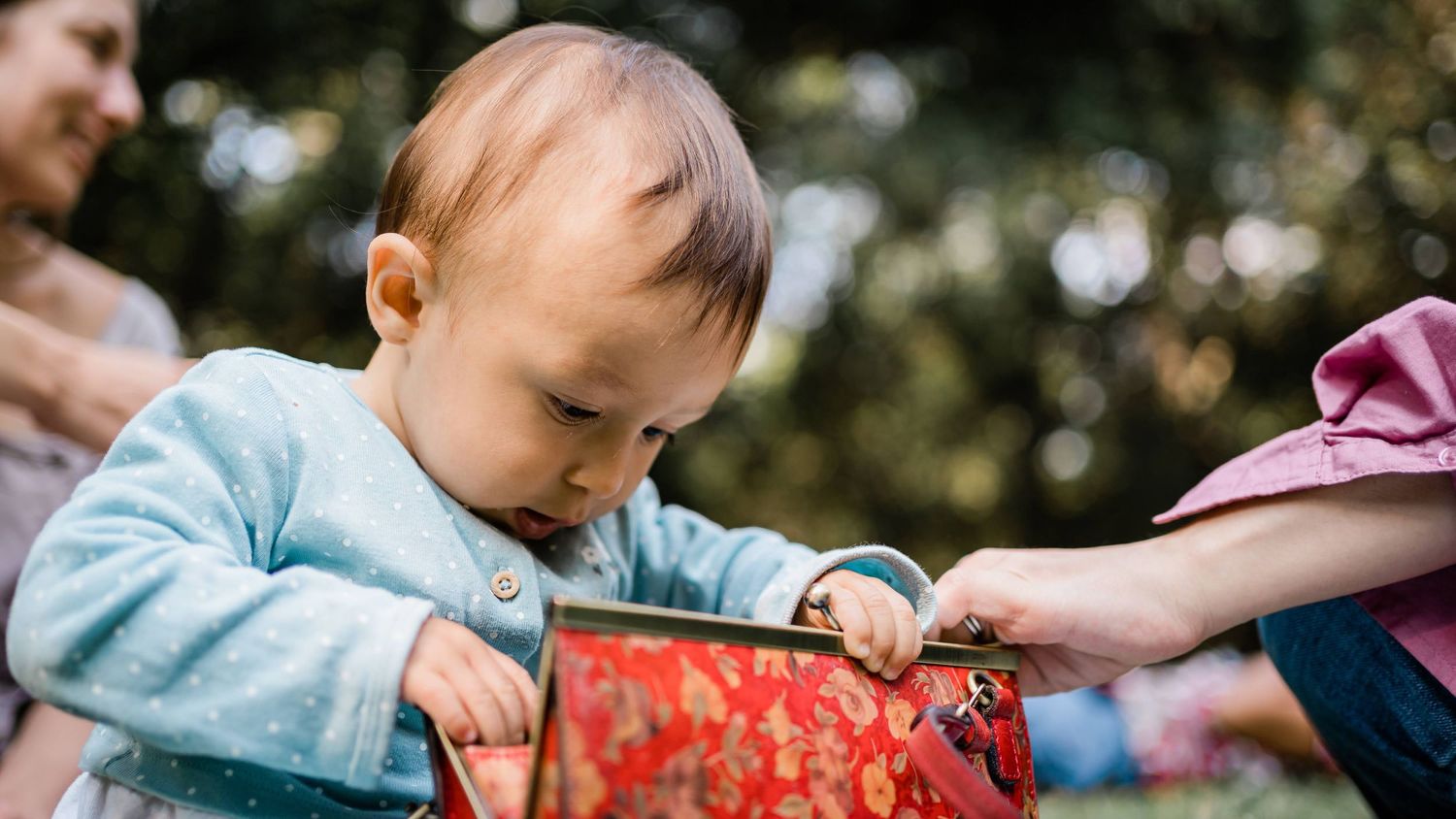 Small child in light blue clothing examines a red purse outdoors.