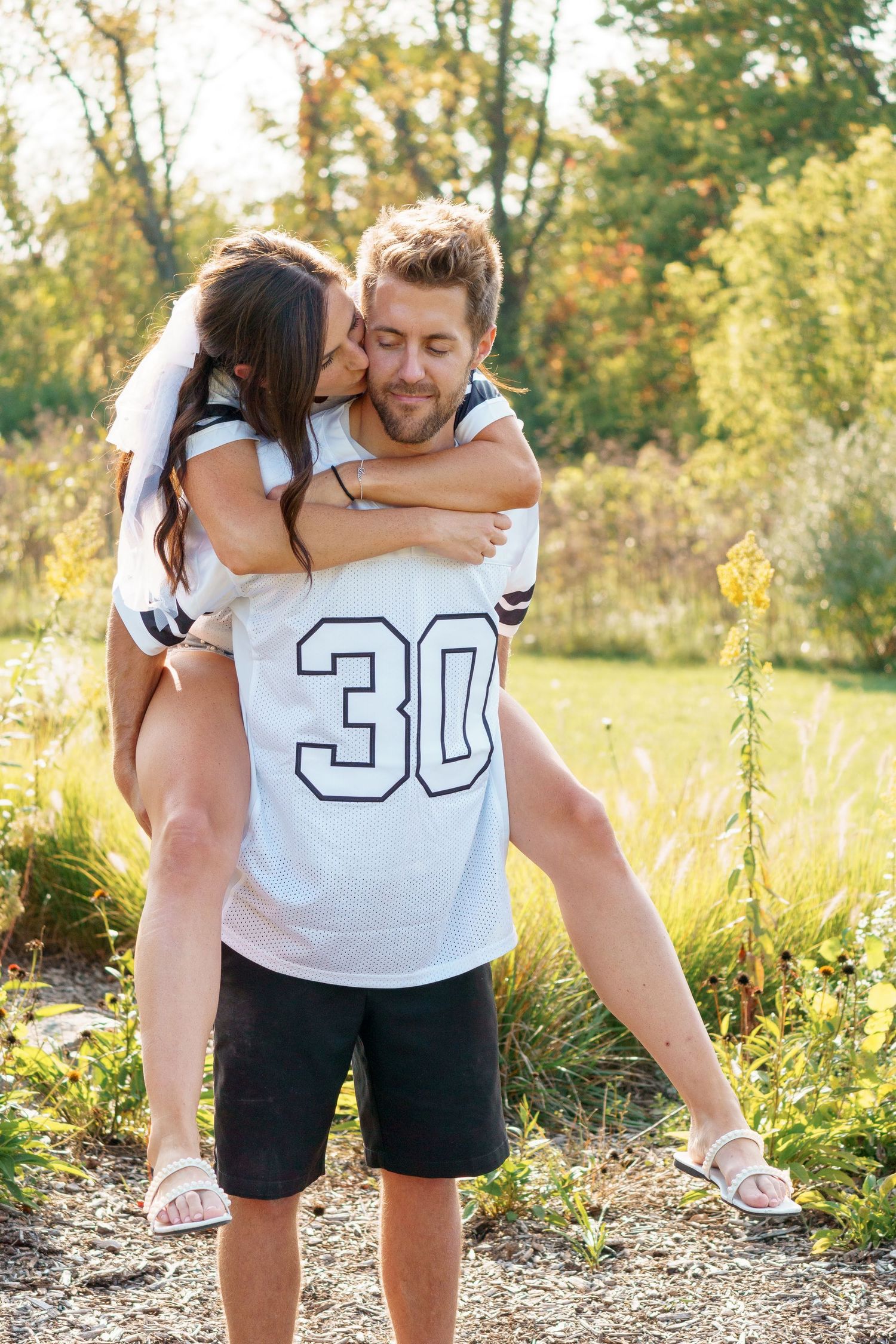 Athletic newlyweds in matching white jerseys sharing romantic moment in nature.