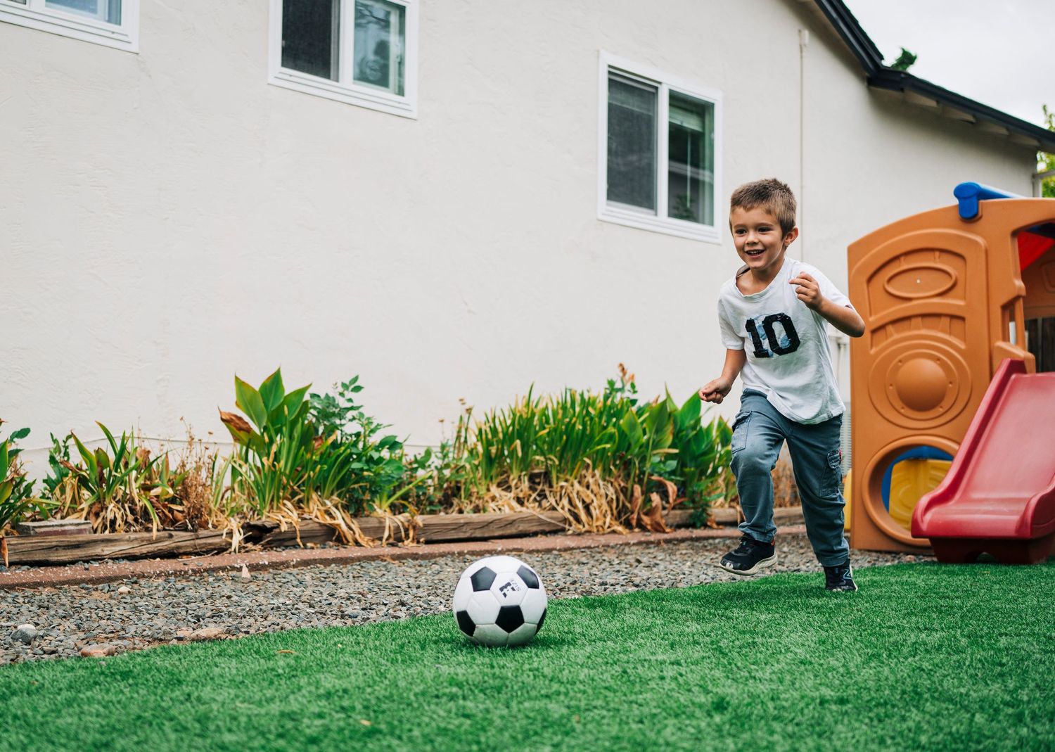 A soccer ball sits on green grass near a plastic toy car and playhouse in a backyard setting.