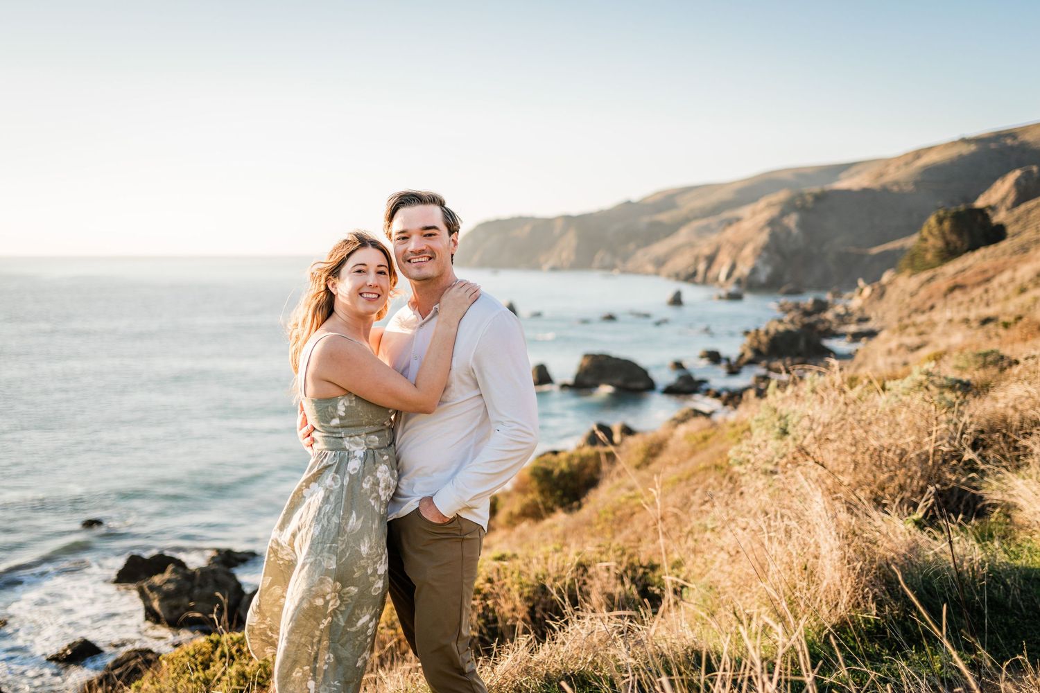 Couple embraces on scenic coastal cliffside during golden hour with ocean waves crashing against rocky shore in background.