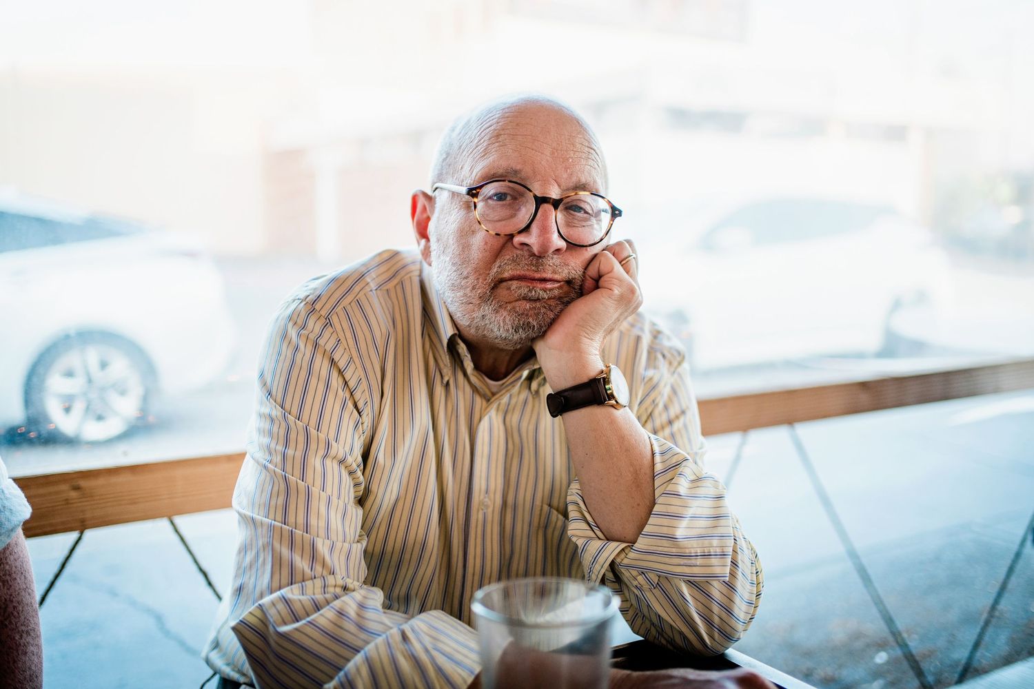 A person in glasses and a striped shirt sits contemplatively at a cafe table with a beverage.