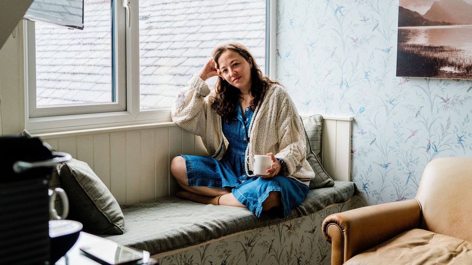 Interior lifestyle scene showing woman drinking tea on couch near white walls and window.