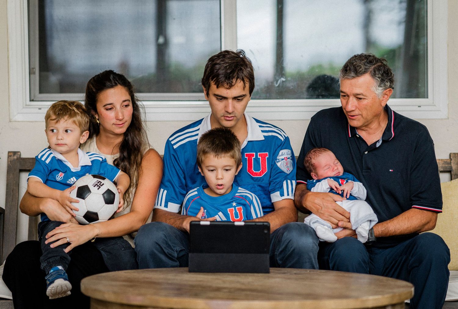 Family gathers around a tablet while watching content together with sports equipment nearby.