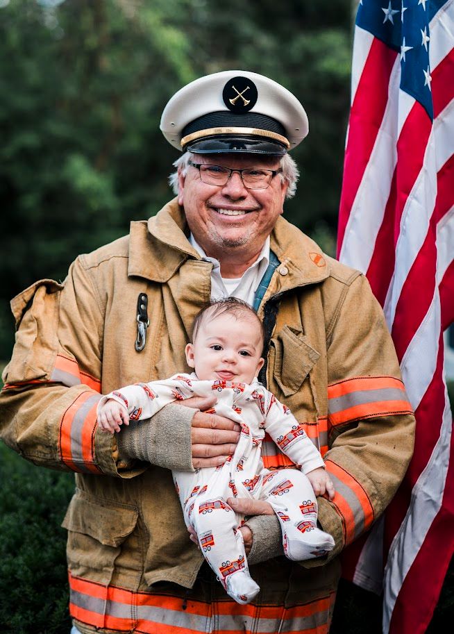 Baby held by firefighter in uniform with American flag backdrop.