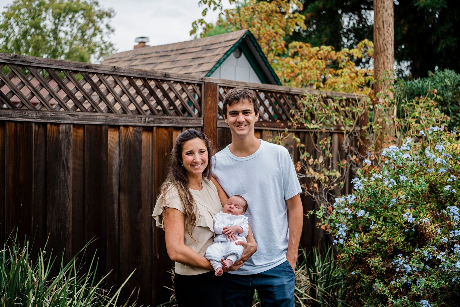 A couple stands together in a garden setting with lush greenery and a wooden fence in the background.
