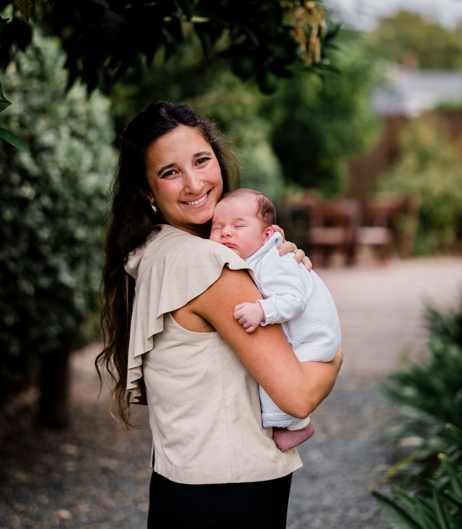 A person in a beige top holds a small baby outdoors among garden greenery.