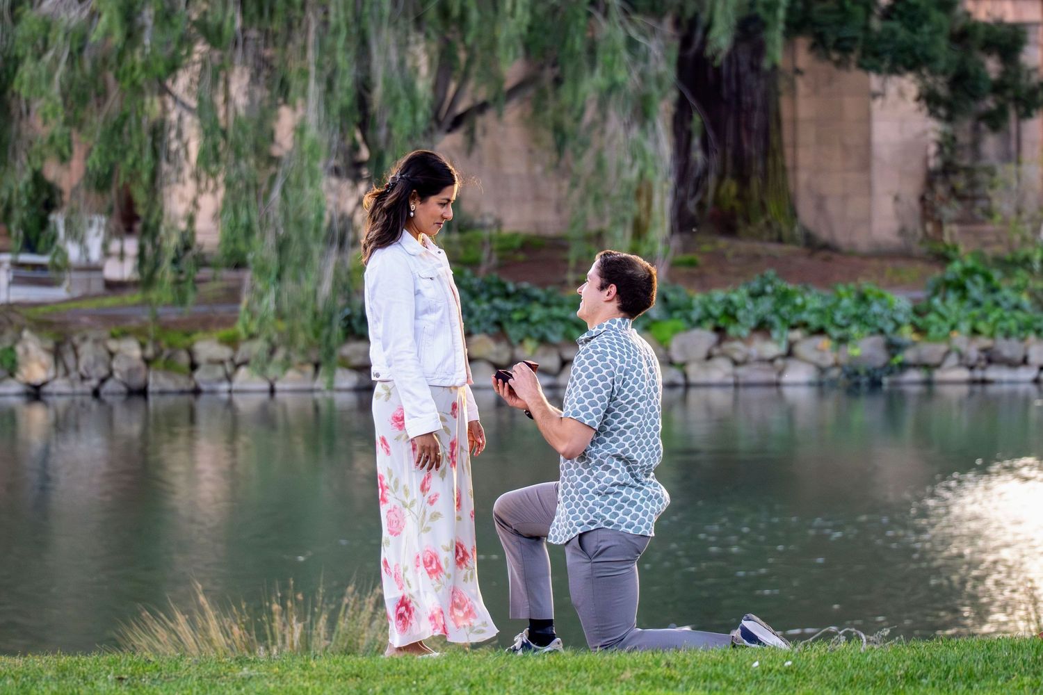 A romantic proposal moment by a peaceful lake with overhanging trees in the background.