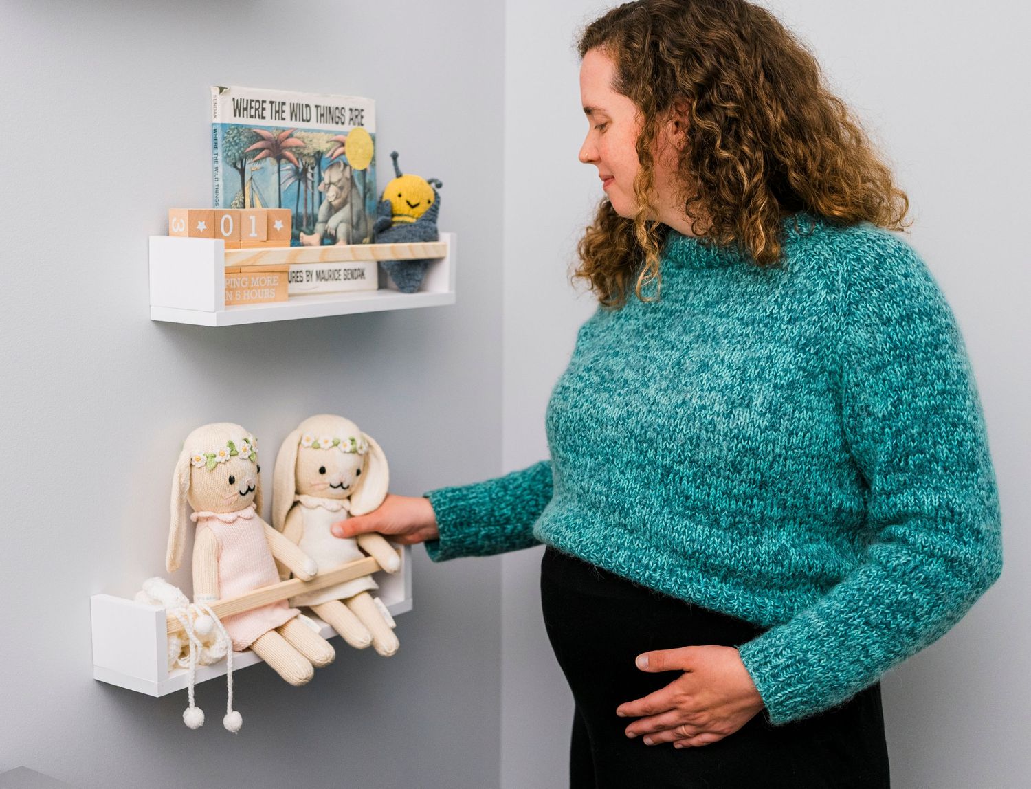 White nursery shelving styled with children's books and plush toys against a minimalist wall with pregnant woman.