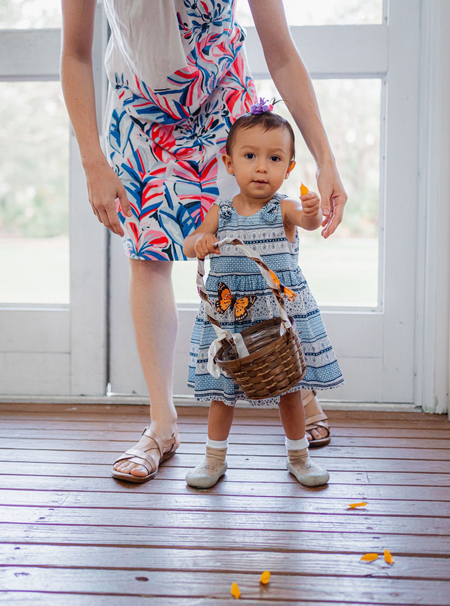 Small child in blue dress stands with caregiver wearing floral dress on wooden porch with scattered flower petals.