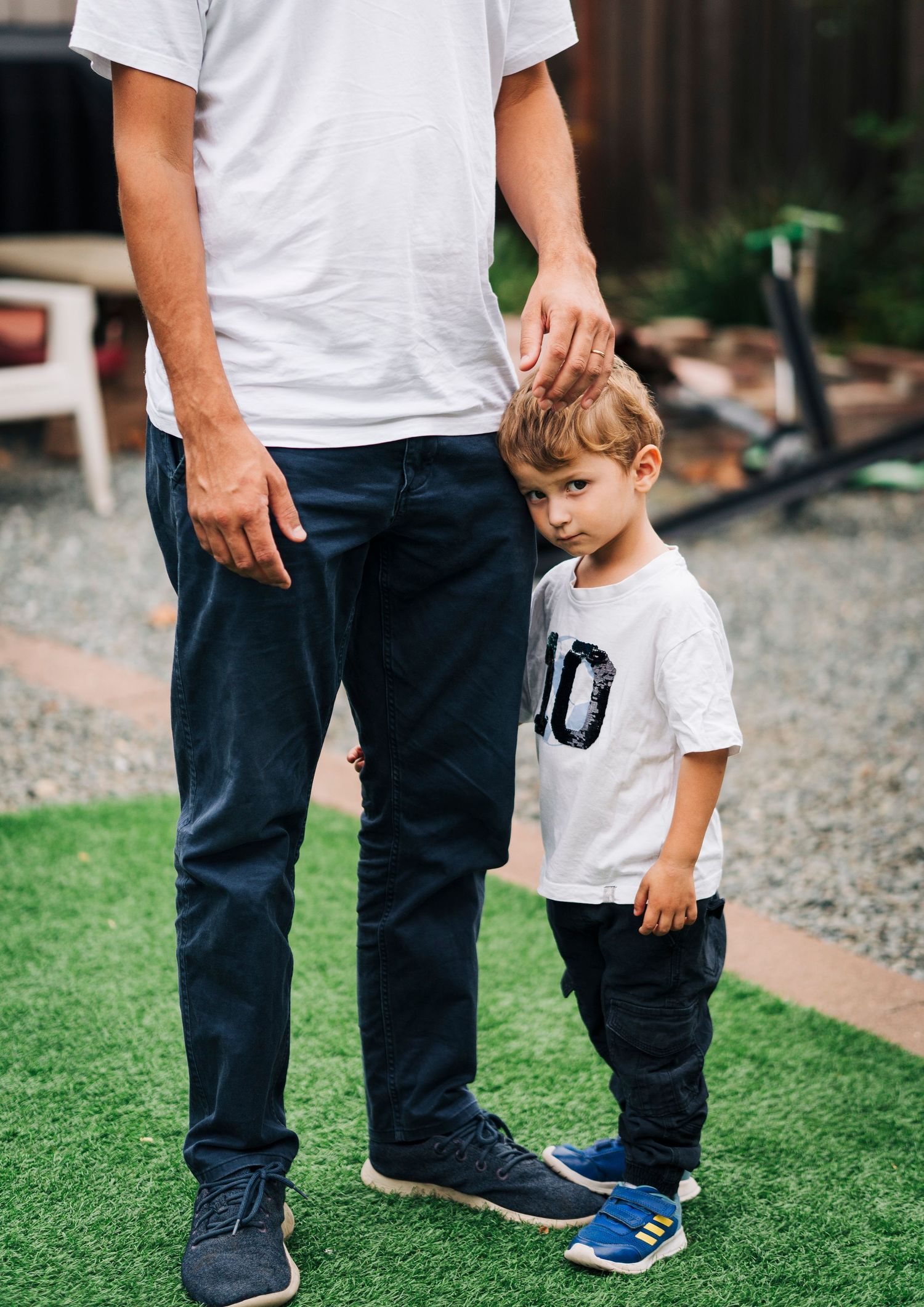 A parent and child stand together on artificial grass, with the child wearing a white t-shirt and blue sneakers.